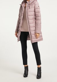 Manteau rembourré rose clair avec fermeture éclair à l'avant et capuche à cordon de serrage, présentant des coutures horizontales. Porté par-dessus un haut beige à côtes et un pantalon noir.