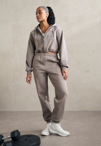 Lichtbruine set bestaande uit een hoodie en joggingsbroek met een cropped ontwerp. Zachte textuur, elastische taille en boorden. Witte sportschoenen.
