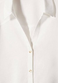 Chemise en lin blanche avec un col plié et une patte de boutonnage ornée de boutons ronds pâles. Texture lisse avec un léger éclat.