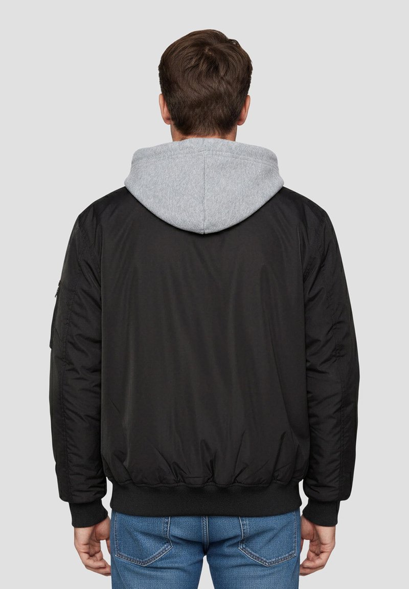 Giacca bomber nera con cappuccio grigio, polsini e vita a coste, tessuto strutturato e tasche laterali. Cuciture visibili sulle maniche.