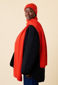 Red ribbed knit beanie and scarf paired with a black quilted coat. The scarf drapes over the shoulders, creating a layered look.
