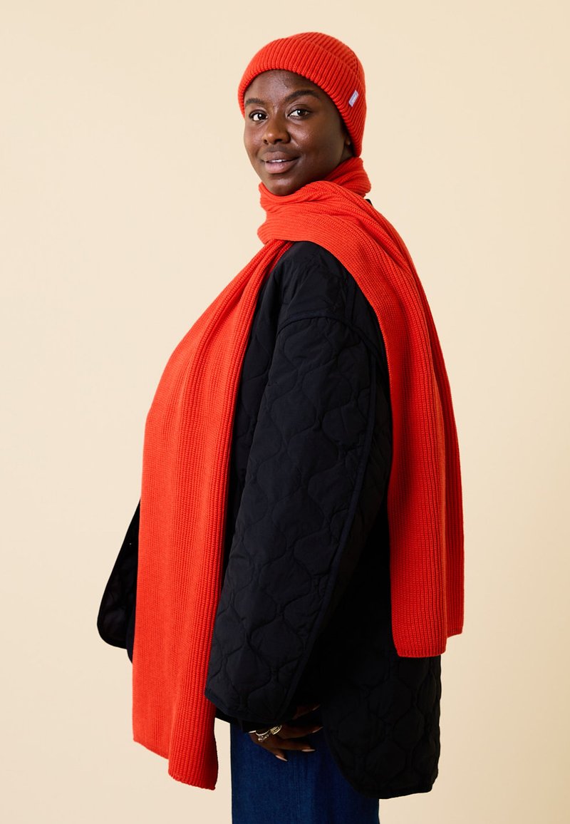 Red ribbed knit beanie and scarf paired with a black quilted coat. The scarf drapes over the shoulders, creating a layered look.