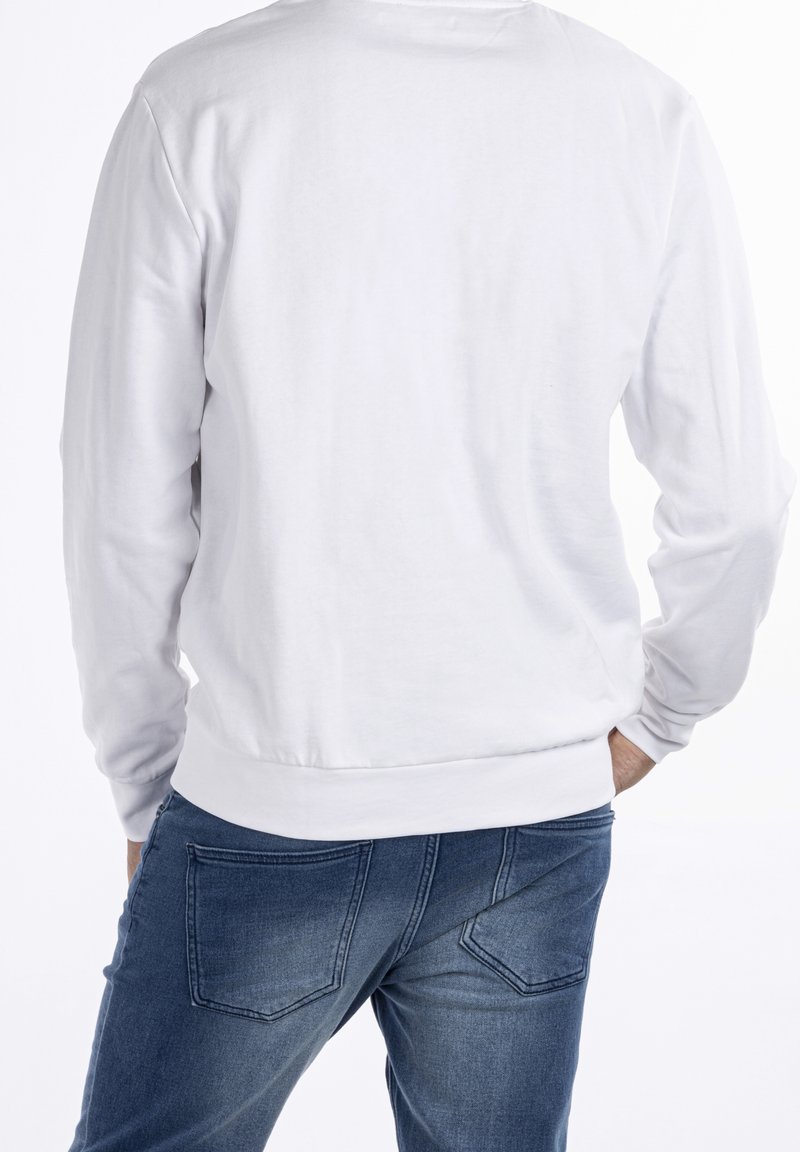 Homme portant une chemise blanche unie à manches longues et un jean bleu en denim, mains partiellement dans les poches arrière, dos tourné à la caméra.