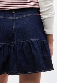 Κοντινή λήψη ενός ατόμου που φοράει μπλε φούστα από τζιν με ζεστές πινελιές και κόκκινη ετικέτα Levi's, σε συνδυασμό με λευκό και κόκκινο ριγέ τοπ.