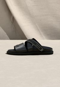 Sandale noire à bout ouvert avec deux larges brides, dont l'une arbore un logo Moschino, sur un fond beige texturé.