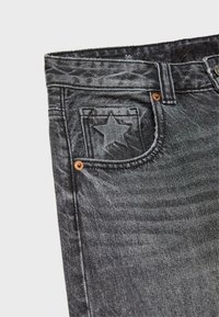 Nahaufnahme von schwarzen Denim-Jeans, die eine Vorderjacke mit sternförmigen Stickdetails und kupferfarbenen Nieten vor einem weißen Hintergrund zeigen.