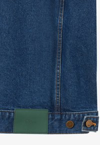 Tissu en denim avec surpiqûres orange, patch en cuir rectangulaire vert, et deux boutons en métal bronze sur le bord d'un jean.