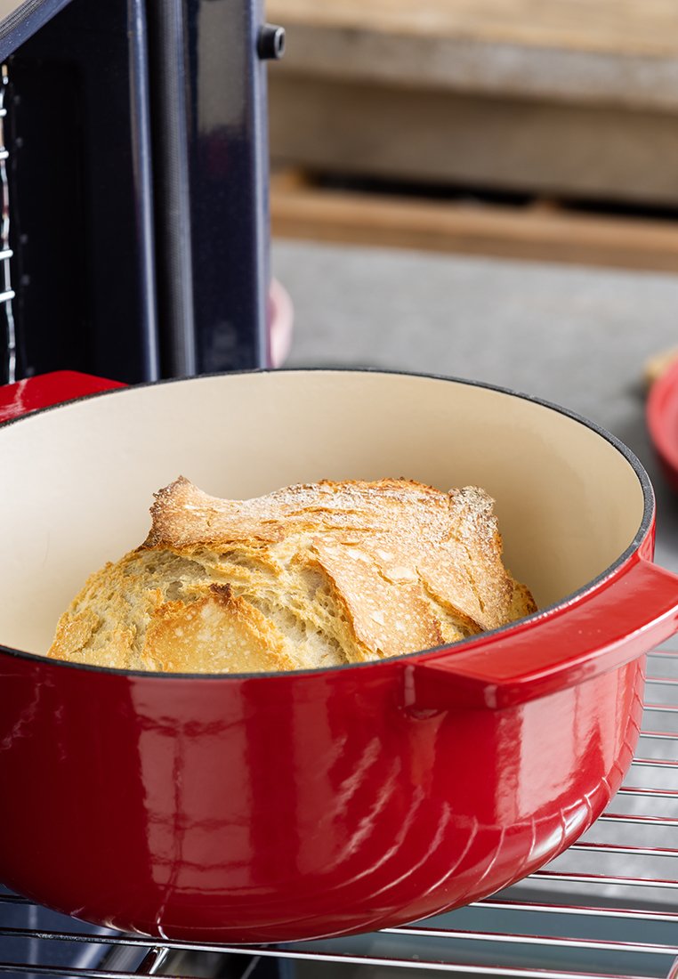 Roter emaillierter Gusseisen-Bräter mit glattem Innenraum, der ein rundes Brot mit goldener Kruste auf einem Drahtgestell enthält.