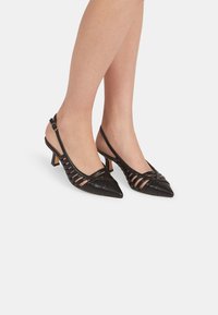 Escarpins slingback en cuir noir avec bout pointu, présentant un design de brides croisées et un petit talon carré.