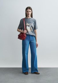 Γυναίκα με γκρι μπλούζα με εκτύπωση αλόγου, μπλε τζιν παντελόνι με φαρδιά μπατζάκια, παπούτσια με εκτύπωση λεοπάρδαλης και κόκκινη τσάντα ώμου.