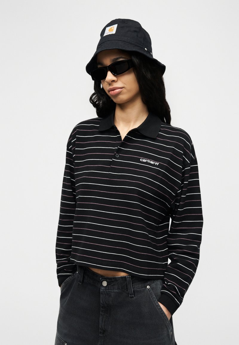 Frau mit schwarzem Fischerhut, schwarzen rechteckigen Sonnenbrillen, gestreiftem schwarzem Langarm-Poloshirt und dunkler, hoch taillierter Jeans, die die Hände in den Taschen hat.