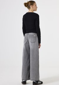 Schwarzes Langarmoberteil mit Rückendetails, kombiniert mit weit geschnittenen grauen Jeans und schwarzen Turnschuhen mit weißen Streifen.