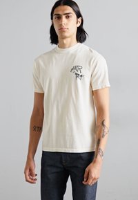 Tricou din bumbac crem cu mâneci scurte, având un imprimeu grafic negru pe partea stângă a pieptului. Asortat cu blugi din denim închis.