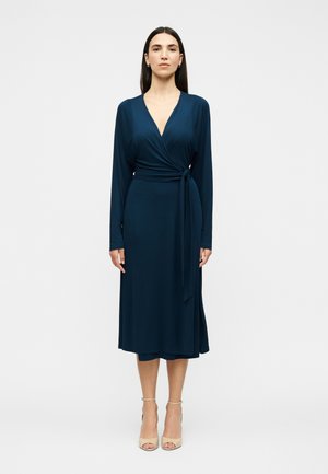 Γυναίκα όρθια, φορώντας μακρυμάνικο μπλε ναυτικό φόρεμα τύπου wrap με ζώνη στη μέση και μπεζ ανοιχτά τακούνια σε λευκό φόντο.