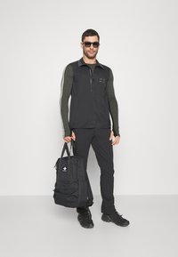 Męska czarna kamizelka na ciemnozielonej koszuli z długim rękawem, czarne spodnie i czarne okulary przeciwsłoneczne, trzymający czarny plecak z logo.