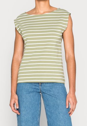 Frau trägt ein ärmelloses, hellgrün-weiß gestreiftes Oberteil mit blauer Jeans, steht vor hellem Hintergrund.