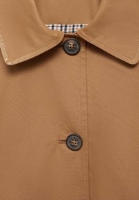 Cappotto in tessuto marrone e liscio con colletto a punta. Presenta una chiusura con bottoni e un dettaglio a motivo check sotto il colletto.
