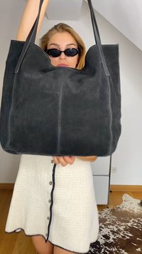 Bolsa tote em camurça preta com alças duplas, apresentando uma textura suave e costura em contraste, segurada por uma pessoa que usa óculos de sol.