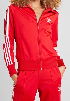 Osoba ubrana w czerwony dres Adidas z białymi paskami na rękawach i spodniach, ręce w kieszeniach oraz srebrny naszyjnik.