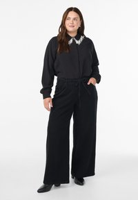 Donna che indossa pantaloni neri a gamba larga e una camicia nera con bottoni e colletto di perline bianche, in piedi con una mano in tasca.
