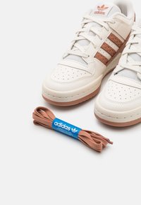 Baskets Adidas blanches et marron avec perforations et bandes texturées, accompagnées de lacets marron supplémentaires dans leur emballage d'origine.