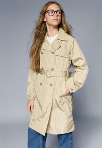 Beige trenchcoat med bælte i taljen, dobbeltknaplukning og elastiske manchetter, lavet af et skinnende, struktureret stof.