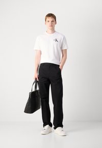 White cotton t-shirt with black "CK" logo, black trousers, white sneakers, and a black tote bag with two handles. Simple, minimal design.