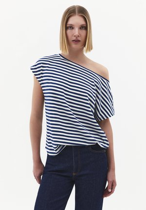 Jonge vrouw met stijl blond haar, een blauw-wit gestreept off-shouldertopje en donkerblauwe spijkerbroek, staand tegen een witte achtergrond.