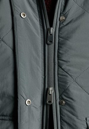 Siva prešita jakna z dvojno zadrgo spredaj, z zaponkami na pritrditev in notranjo teksturirano podlogo. Vidna so teksturirano blago in detajli šivov.