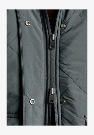 Siva prešita jakna z dvojno zadrgo spredaj, z zaponkami na pritrditev in notranjo teksturirano podlogo. Vidna so teksturirano blago in detajli šivov.