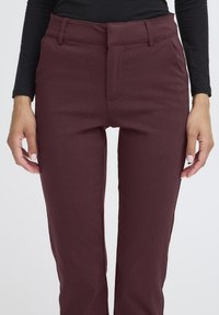 Bordeauxkleurige broek van rekbaar materiaal, met een vlakke voorkant, ceintuurlussen en een getailleerde pasvorm, getoond vanaf de taille naar beneden.