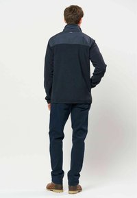 Homme debout, de dos, portant une veste bleu marine, un pantalon bleu foncé et des bottes marron sur un fond clair uni.