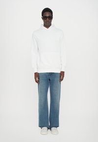 White hooded sweatshirt with a front pocket, blue wide-leg jeans, and white sneakers. Dark sunglasses complete the casual look.