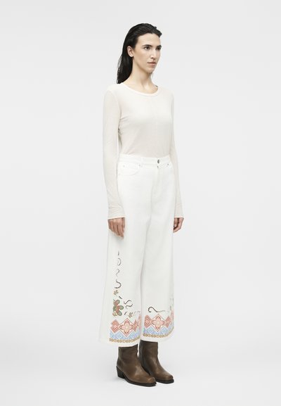 Γυναίκα με μακριά σκούρα μαλλιά που φοράει διάφανο μπεζ μακρυμάνικο τοπ, λευκό παντελόνι φαρδιάς γραμμής με πολύχρωμη κεντημένη ανθοδέσμη και καφέ μποτάκια αστραγάλου.