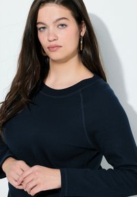 Marineblauw, langärmeliges Oberteil mit einer strukturierten Rippenoptik, rundem Halsausschnitt und genähten Details an den Ärmeln. Das Modell hält die Hände vor sich.