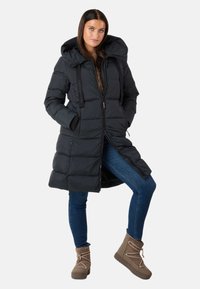Giacca a vento nera con cappuccio, frontale con zip e stringhe. Presenta un design trapuntato e tasche laterali, abbinata a jeans blu e stivali marroni.