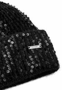 Czarna, dziana czapka ozdobiona płaskimi, czarnymi cekinami. Posiada srebrną metkę z grawerem, ukazującą teksturowaną i refleksyjną powierzchnię.