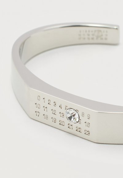 Ασημένιο βραχιόλι μανσέτα από μέταλλο με χαραγμένους αριθμούς από το 0 έως το 23 και μία διάφανη κρυστάλλινη πέτρα στο νούμερο 21, σε λευκό φόντο.