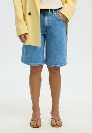 Person trägt kniehohe blaue Jeansshorts, gelben Blazer, neutrales Top, braunen Gürtel und sandfarbene offene Sandalen, steht mit einer Hand in der Tasche.