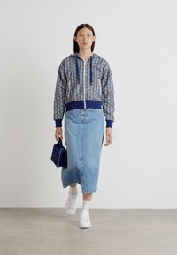 Hanorac cu fermoar, cu un model geometric în nuanțe de bleumarin și gri, purtat cu o fustă din denim de lungime medie și adidași albi, ținând în mână o geantă mică albastră.