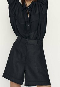 Chemise noire surdimensionnée avec un nœud à l'avant et deux poches poitrine, associée à un short noir taille haute à jambes larges. Le tout est fabriqué dans un tissu léger.