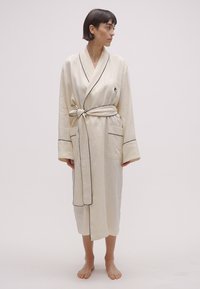 Gaiši bēša halāts ar melnām maliņām, gara piedurkne, ar piesaistes jostiņu un kabatām, izgatavots no gludas auduma, ceļgalu garumā.