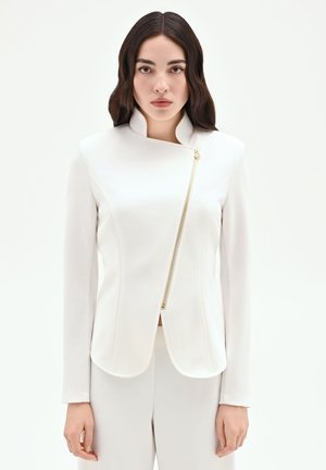 Femme aux longs cheveux foncés porte une veste blanche ajustée avec une fermeture éclair dorée asymétrique et un pantalon blanc assorti, debout devant un fond blanc.