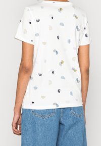Camiseta blanca de manga corta con un patrón abstracto de puntos multicolor. La tela tiene una textura suave; corte relajado con un cuello redondo.