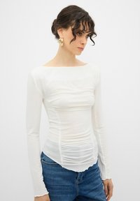 Vit långärmad topp med samlad textur och båtformad halsringning, tillsammans med blå denimjeans. Slät tygkvalitet, välsittande silhuett.