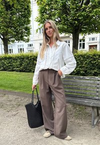 White blouse with lace detailing, brown wide-leg pants, and a black woven tote bag. Standing on a path near a bench and greenery.