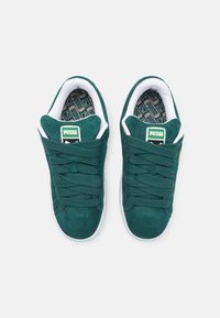 Gröna mockaskor med en vit gummisula, med texturerade snörningar och ett präglat mönster på innersulan, märkt med en Puma-logga.