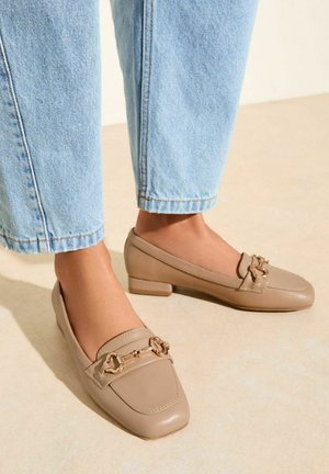 Mocassins en cuir beige avec un bout carré et un petit talon bloc, ornés d'une chaîne dorée sur le devant. Portés avec un jean en denim bleu clair.