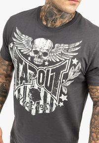 Graues Baumwoll-T-Shirt mit einem verwaschenen weißen Totenschädel und Flügelgrafik, sowie dem Text "TAPOUT" und "seit 1991" in fetter Schrift.