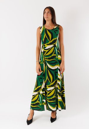 Abito maxi verde con grandi motivi a foglia gialla e bianca. Presenta un collo rotondo e una silhouette fluida. Scarpe nere con tacco completano l'outfit.